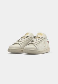 Kaks off-white värvi Adidas Stan Smithi tenniset, millel on beežid tallad, kuldne bränding keelel ja külgedel ning mitmevärvilised kontsaplaadi aktsendid.