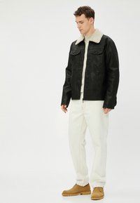 Zwarte leren jas met een crème faux schapenvacht kraag, voorzakken en rits. Gecombineerd met witte broek en beige schoenen.
