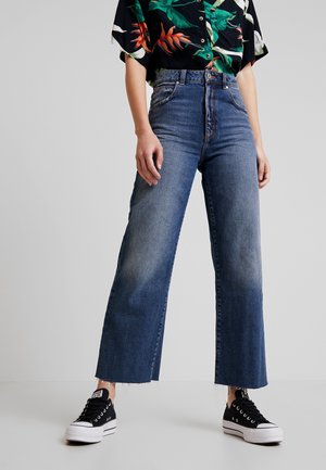 Person iført blå jeans med høj talje og vide ben med rå kanter, sorte og hvide sneakers og en sort skjorte med farverigt tropisk print.