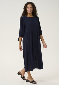 Vestido azul marino hecho de tejido ligero, con un diseño en capas, mangas abullonadas de tres cuartos y detalles de lazo en los puños.