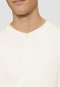 Valge henley-tüüpi särk, millel on tekstuurne kangas, kolm nööpi nööpplaadil ja V-kujuline kaelus.