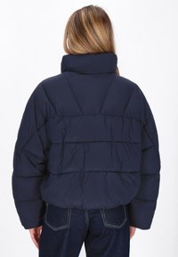 Jachetă puffer de culoare bleumarin, cu guler înalt, cusături orizontale și croială lejeră. Textura moale, matlasată, vizibilă din spate.