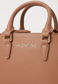 Tan värvi kott, mis on valmistatud tekstureeritud materjalist, kahe käepideme ja kuldse viimistlusega. Kaubamärgi nimi "VALENTINO" on graveeritud sujuvas fondis.