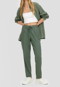 Grüne Leinenhose mit Kordelzugbund und vorderen Falten, kombiniert mit einem weißen Cropped-Top und einem übergroßen grünen Hemd mit Knöpfen.