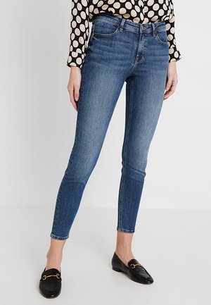 Kobieta w niebieskich, obcisłych jeansach z podwyższonym stanem, czarnych loafersach oraz czarno-beżowej bluzce w grochy z długim rękawem, stojąca na neutralnym tle.