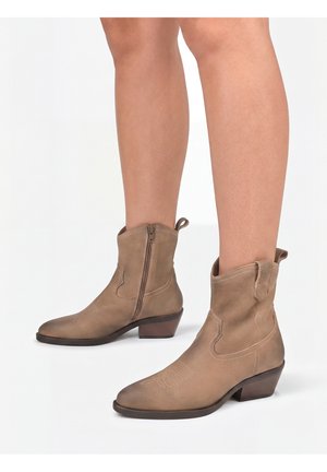 Brune ruskind ankleboots med lynlåse i siden og lav blokhæl båret på bare ben mod en hvid baggrund.