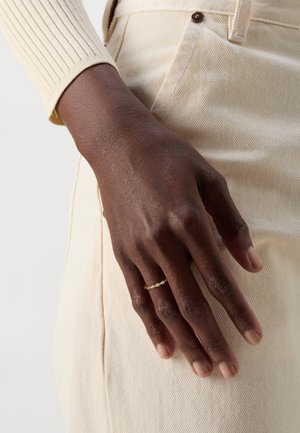 Gouden ring met kleine ronde accenten, gedragen op een hand met een donkere huidtint. Hand ligt op een lichtbeige stof met een gestructureerd oppervlak.