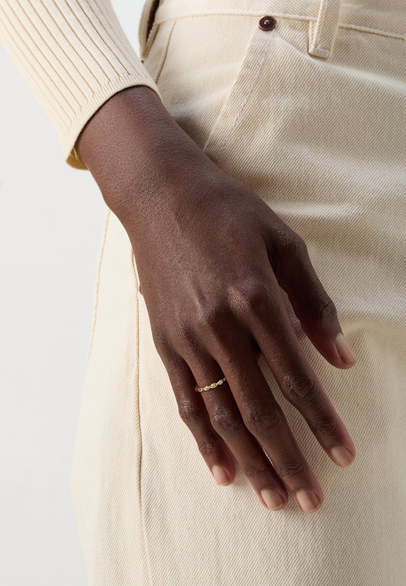 Bague en or avec de petits accents ronds portée sur une main à la peau foncée. La main repose sur un tissu beige clair avec une surface texturée.