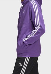 Lila hoodie med vita ränder på ärmarna, justerbar dragsko på huvan, sidofickor, i kombination med svarta byxor med vita ränder och logotyp.