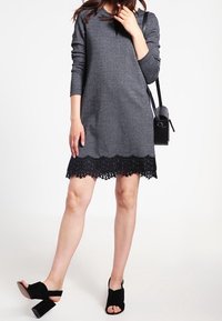 Szara dzianinowa sukienka z długimi rękawami i czarnym koronkowym wykończeniem. Modelka ma na sobie czarne sandały na grubym obcasie i trzyma małą czarną torebkę.