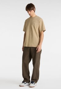 Beige cotton t-shirt with short sleeves, worn with brown denim pants. The outfit is completed with white sneakers and navy laces.