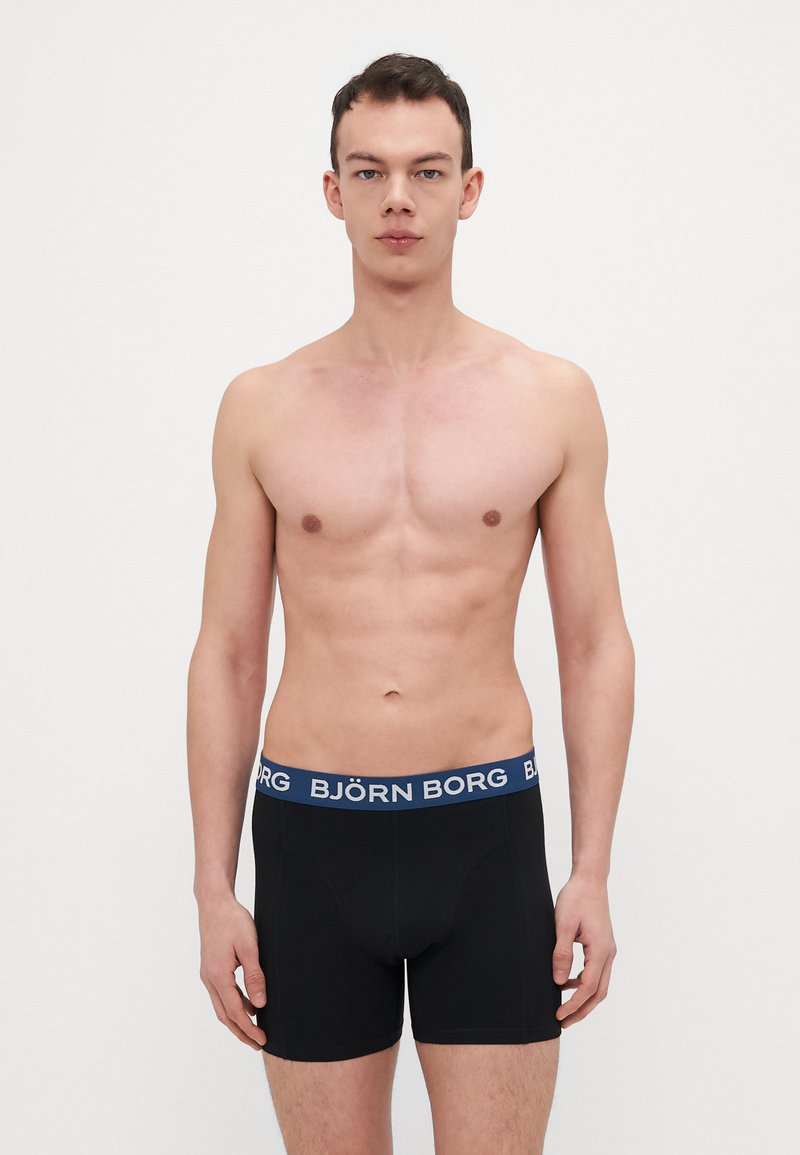 Svarta boxershorts med blå elastisk midjeband som har "BJÖRN BORG"-logotyp; figurnära design, slät textur och längd till mitten av låret.