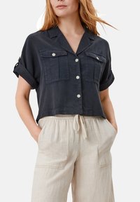 Camicia corta navy in tessuto morbido con colletto, due tasche frontali e chiusura a bottoni. Abbinata a pantaloni larghi color beige chiaro.