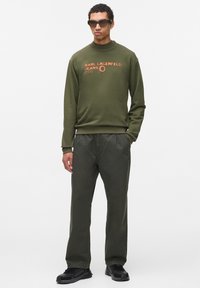 Sudadera verde oliva con texto gráfico, combinada con pantalones verdes oscuros. Los accesorios incluyen gafas de sol negras y zapatillas. Tejido suave y casual.