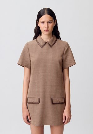 Modella che indossa un vestito corto a scacchi marrone con colletto e due tasche frontali, in piedi contro uno sfondo semplice, con un'espressione neutra.