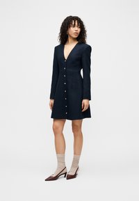 Γυναίκα με σγουρά μαλλιά που φοράει μακρυμάνικο μπλε φόρεμα με κουμπιά μπροστά, μπεζ κάλτσες και καφέ χαμηλά παπούτσια με μυτερό άκρο, στέκεται σε λευκό φόντο.