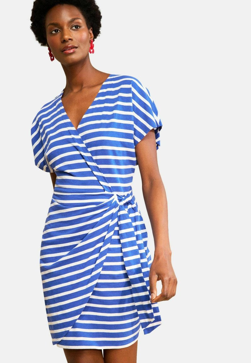 Femme portant une robe portefeuille à rayures horizontales bleues et blanches avec des manches courtes et des boucles d'oreilles rouges en forme d'anneaux.