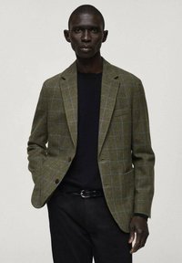 Blazer a quadri verdi e blu con design strutturato, dotato di due bottoni e rever a punta, indossato sopra un maglione nero.