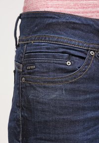 Tmavě modré džíny z denimu s pětipocketovým designem, kontrastním prošíváním a malým kovovým logem na zadní kapse.