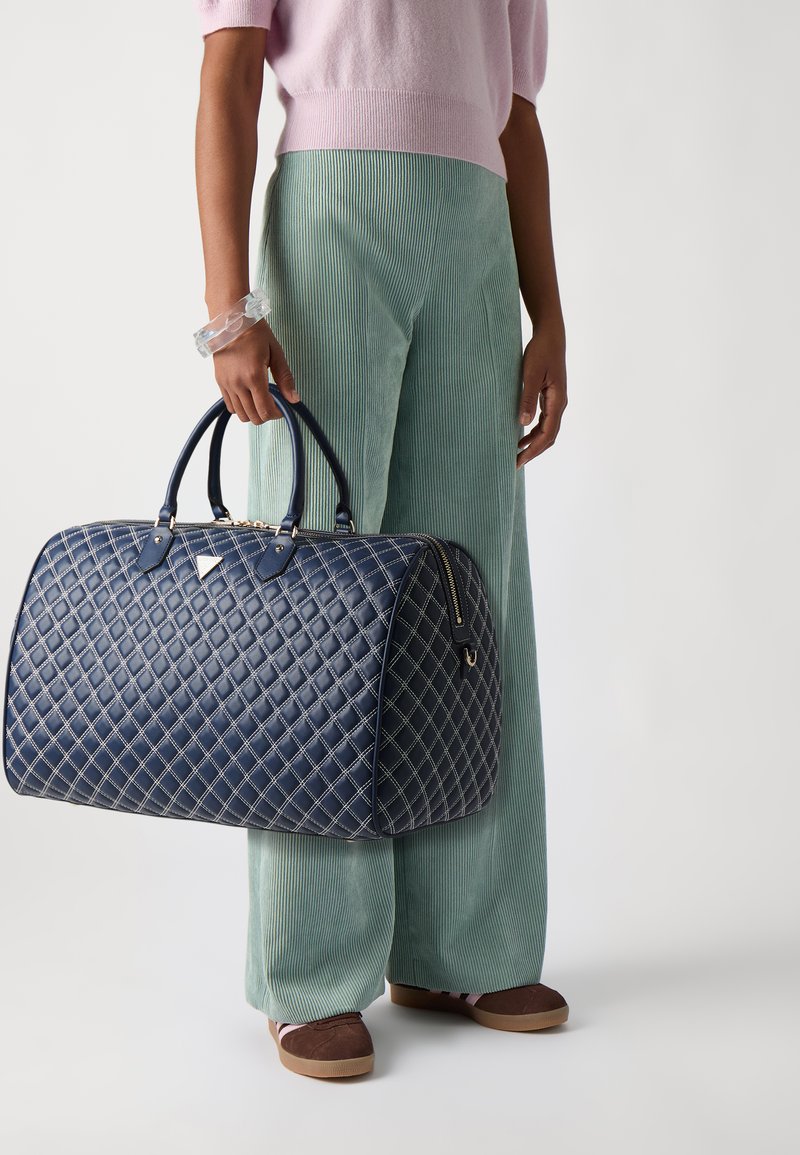 Borsa da viaggio in pelle blu navy trapuntata con manici doppi, dettagli in metallo dorato e cuciture crema. Il modello indossa pantaloni in corduroy verde chiaro.