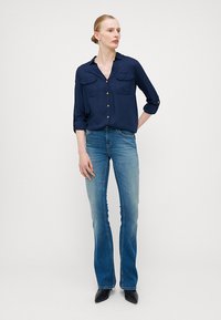 Chemise boutonnée bleu marine avec deux poches poitrine, associée à un jean flare bleu clair. L'ensemble est complété par des chaussures à talons noires.