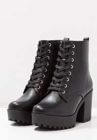 Botas de tobillo de cuero negro con un tacón alto de bloque, punta redonda y cordones en la parte frontal. Presenta ojales de metal y una suela gruesa con dibujo.
