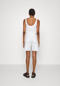 Mujer con cabello trenzado, vistiendo una blusa blanca sin mangas y pantalones cortos blancos hasta la rodilla, con sandalias negras, de pie frente a un fondo liso.