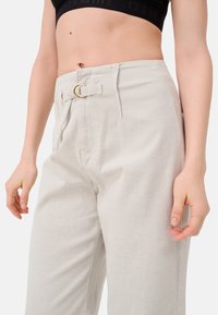Lichtgrijze broek van gestructureerde stof, met een goudkleurige riemgesp en zijzakken. Hoog in de taille.