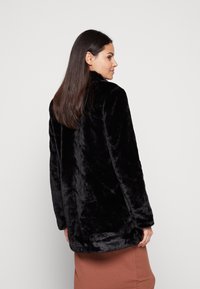 Manteau en fausse fourrure noire avec un col montant, coupe décontractée et texture lisse. La vue de dos montre des manches longues et un drapé doux.