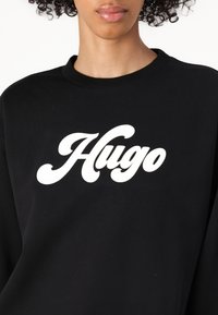 Felpa nera realizzata in tessuto morbido, caratterizzata da un logo bianco "Hugo" in uno stile corsivo arrotondato. Design classico con scollo rotondo.
