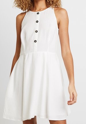 Ærmeløs hvid kjole med rund halsudskæring og fire sorte knapper foran, båret af en person med krøllet hår.