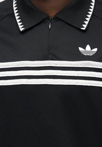 Μαύρο πόλο μπλουζάκι με φερμουάρ στο γιακά, λευκές οριζόντιες ρίγες και χαρακτηριστικό ζιγκ-ζαγκ μοτίβο στον γιακά. Λογότυπο Adidas στο στήθος.