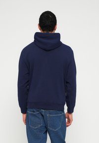 Tmavomodrá mikina s veľkou, mäkkou kapucňou, rebrovanými manžetami a lemom; nosená s modrými džínsami, pohľad zozadu.