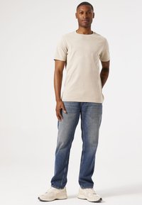 Beige Baumwoll-T-Shirt mit kurzen Ärmeln, kombiniert mit blauen Jeans und hellen Sportschuhen, vor neutralem Hintergrund stehend.