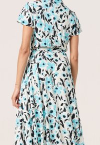 Květinové šaty s lehce modrým a černým abstraktním vzorem na bílém pozadí, vyrobené z měkké látky, s přiléhajícím pasem a rozšířenou sukní.