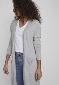 Ljusgrå cardigan i mjukt material, med ribbade manschetter, öppen front och två framfickor; bärs över en vit T-shirt med V-hals.