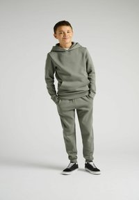 Ensemble de jogging vert composé d'un sweat à capuche avec une poche frontale et d'un pantalon fuselé assorti, associé à des baskets noires.