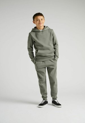 Grünes Jogging-Set mit einem kapuzenpulli mit einer Fronttasche und passenden schmal geschnittenen Hosen, kombiniert mit schwarzen Sneakers.