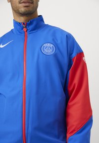 Blue and red Nike jacket with a stand-up collar, featuring a front zipper, side pockets, and a circular logo on the chest.