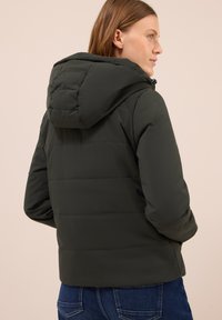 Veste matelassée verte foncée avec capuche, dotée d'un design matelassé et de poignets élastiques. Texture lisse. Portée sur un jean en denim.