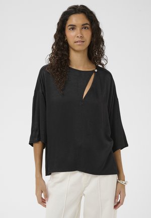 Femeie cu păr creț purtând un top negru larg cu o mică decupare și pantaloni albi, pe un fundal simplu și deschis la culoare.