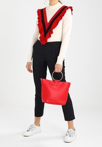 Rød mulepose med cirkulære håndtag, glat tekstur. Bruges med en hvid sweater med rødt og mørkeblåt flæsemønster i V-form og bukser med nålestriber.