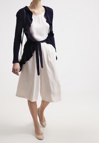 Námořnicky modrý kardigan s volánky přes bílou blůzu, doplněný krémovými širokými kalhotami a béžovými balerínami, s uvázaným pasem a hladkou texturou.