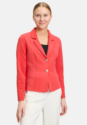 Blazer rosso su misura con colletto a punta, due bottoni e design aderente. Maniche lunghe e tessuto liscio. Indossato sopra un top nero.