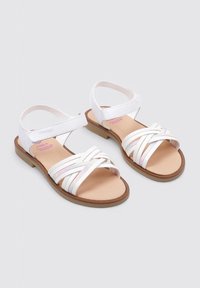 Sandalias blancas con correas entrelazadas, que presentan un acento brillante. Suela plana de goma marrón y correas ajustables en el tobillo para mayor comodidad.