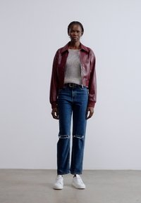 Bordó bőrbomber dzseki, világosszürke kötött pulóver, kopott kék farmernadrág és fehér tornacipők. A modell semleges háttér előtt áll.