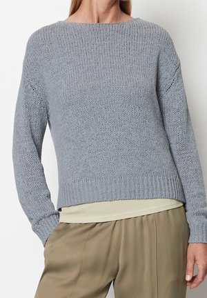 Kvinde iført en lys grå strikket sweater over en cremefarvet top med beige bukser, stående med den ene hånd i lommen mod en ensfarvet baggrund.