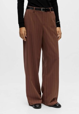 Bruine wijde pantalons met krijtstreep, gecombineerd met een zwarte riem en zwarte schoenen, gedragen met een zwart shirt met lange mouwen, zicht op het onderlichaam.