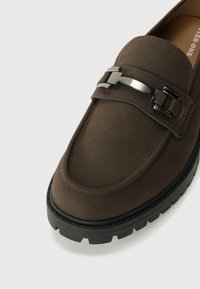 Mocasín de ante marrón con un acento de hebilla plateada, que presenta una gruesa suela de goma negra y un diseño de punta redondeada y limpio.