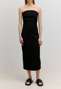 Schwarzes trägerloses Kleid aus glattem Stoff, figurbetontes Design, knielang mit subtilen Rüschen; getragen mit schwarzen Riemchensandalen.